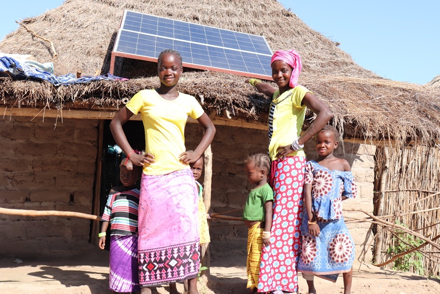 Two women stand outside their home, which has a thatched roof and solar panel on that roof. Four children stand around the women's legs.