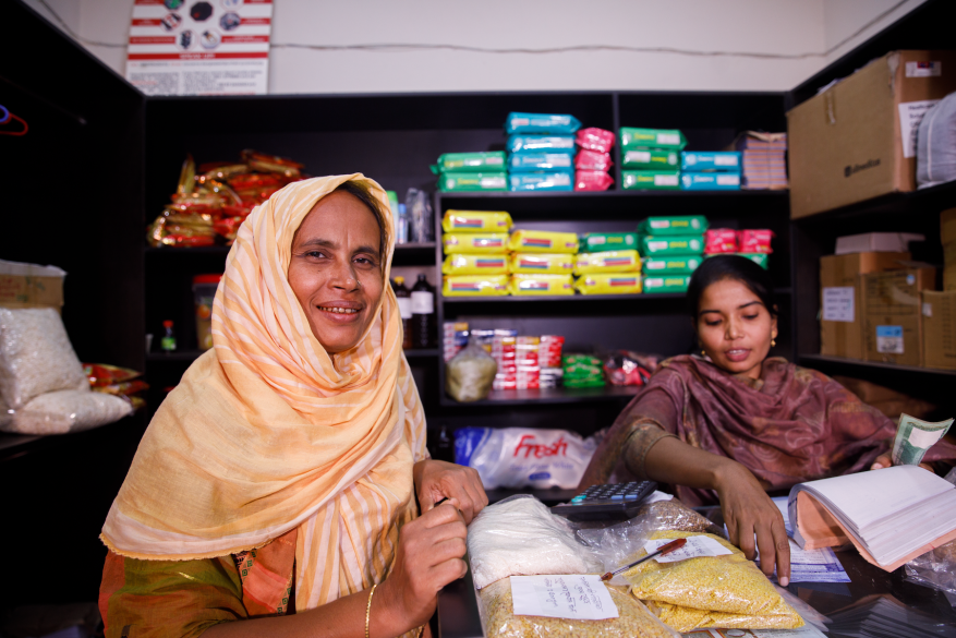 Two rural women entrepreneurs in Bangladesh stand at a shop desk with shelves behind them. The shelves are stacked with colourful packets.
