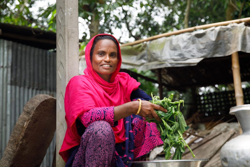 Woman in pink shawl covering head and shoulders sits outside smiling and holding green vegetables.