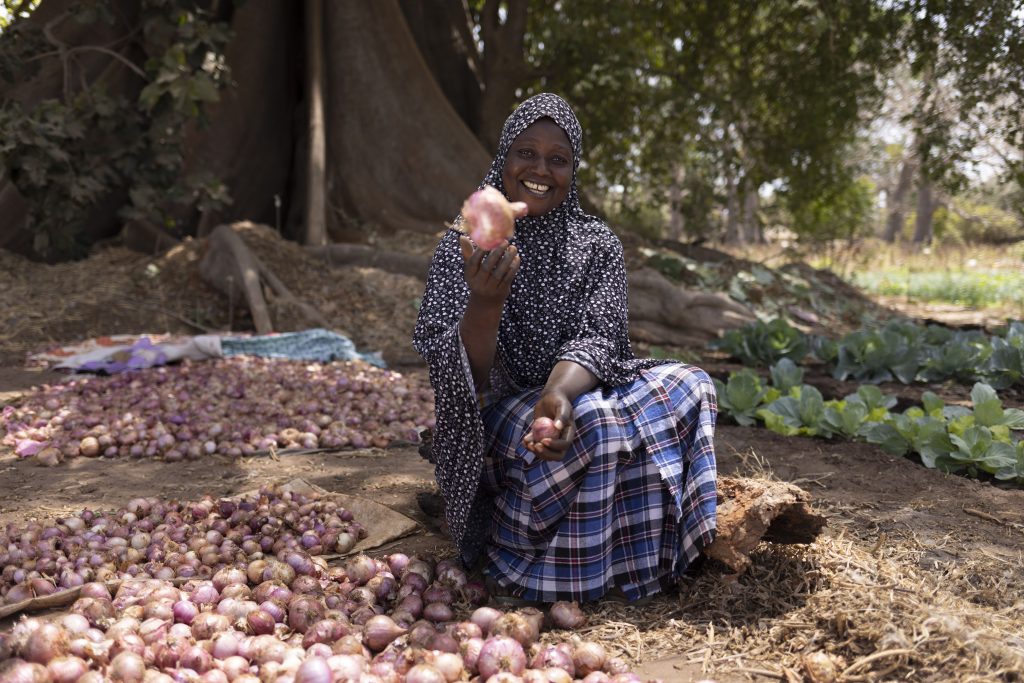 Woman farmer sits among harvested onions, smiling and holding one onion up to the camera.