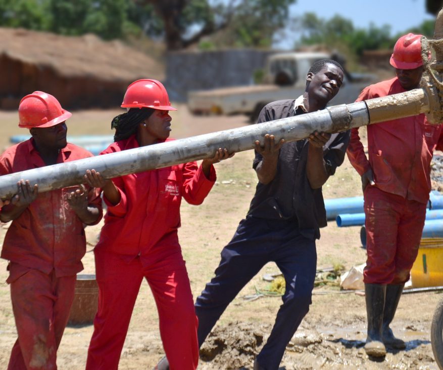 Four people - three of whom are dressed in red overalls and wearing helmets - at work drilling a well.