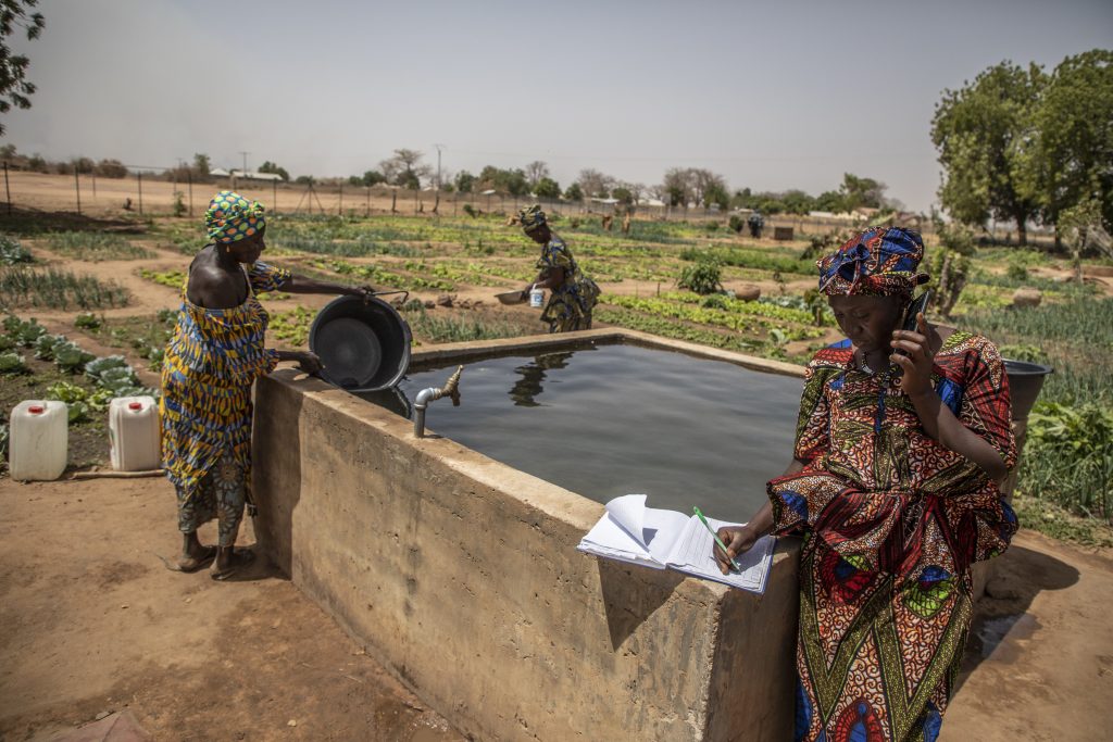 Woman  in foreground of photo sits near water writing on paper while holding phone. Other women use water in background.